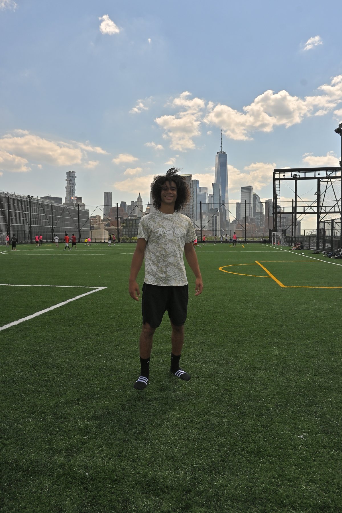 Davide Clarkson wearing a JOGA NYC shirt on a soccer field with the New York City skyline behind him